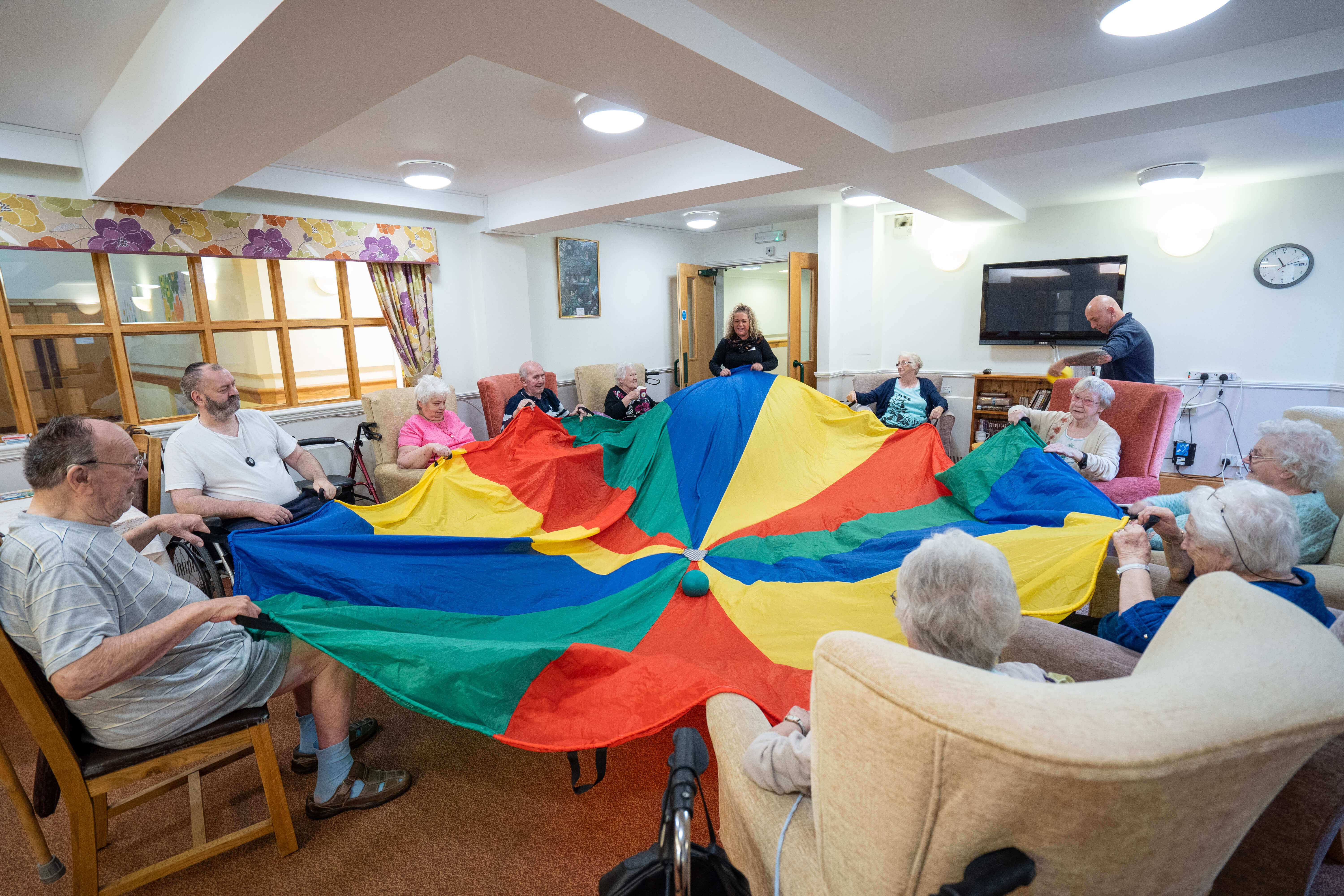 Older people sitting on couches holding a blue, green, red and yellow stripped cloth rolling a green ball in the middle on the cloth to one another
