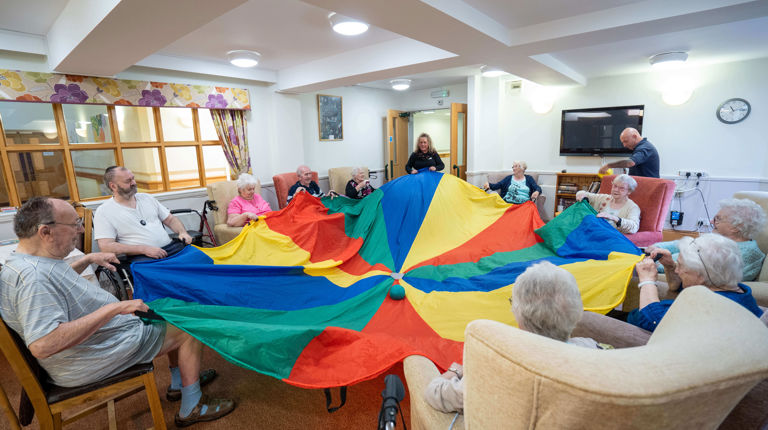 Older people sitting on couches holding a blue, green, red and yellow stripped cloth rolling a green ball in the middle on the cloth to one another Older people sitting on couches holding a blue, green, red and yellow stripped cloth rolling a green ball in the middle on the cloth to one another
