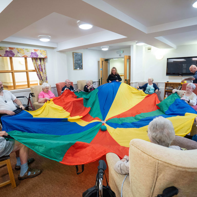 Older people sitting on couches holding a blue, green, red and yellow stripped cloth rolling a green ball in the middle on the cloth to one another Older people sitting on couches holding a blue, green, red and yellow stripped cloth rolling a green ball in the middle on the cloth to one another