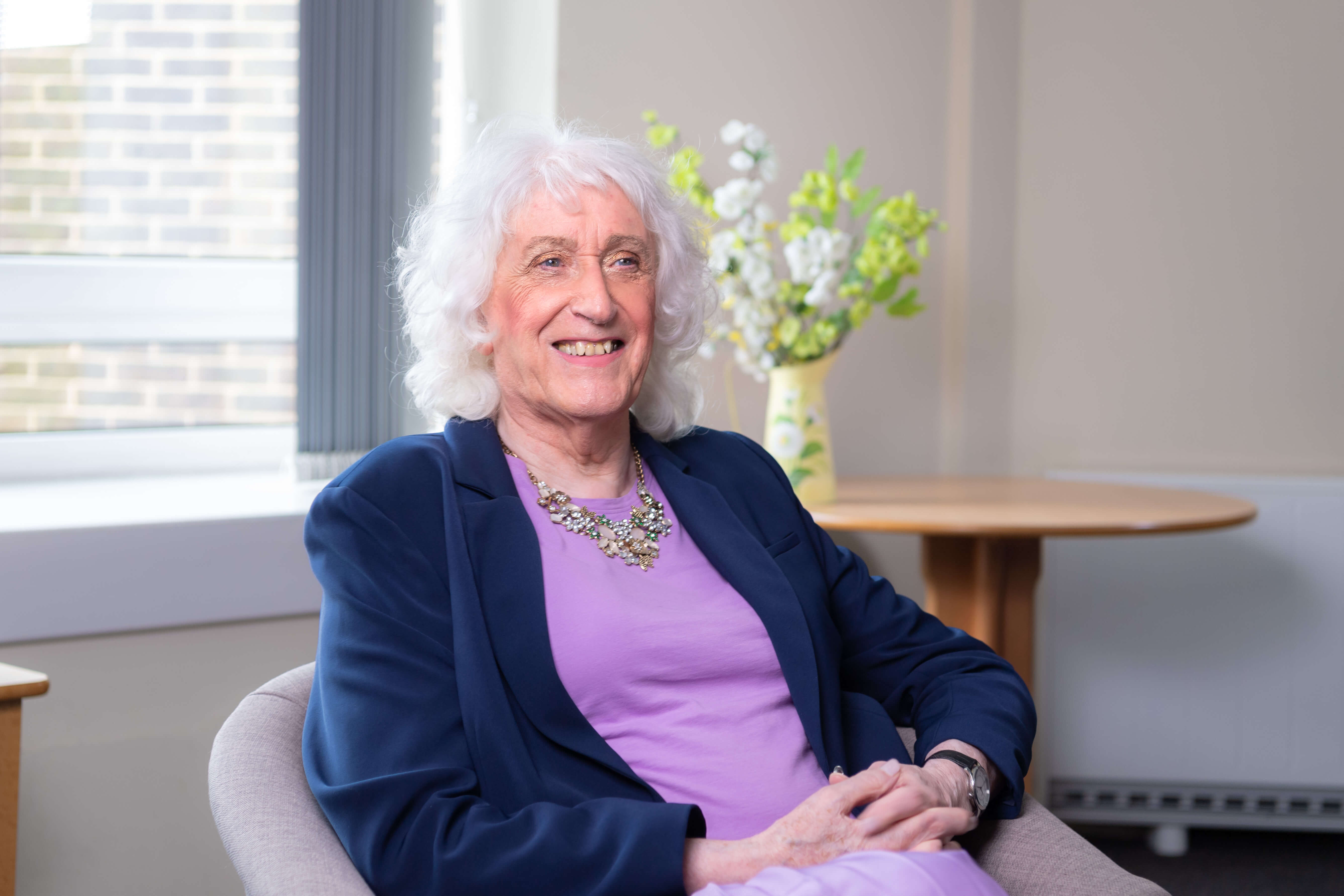 Sandra sitting on a couch wearing a navy blue blazer and purple dress