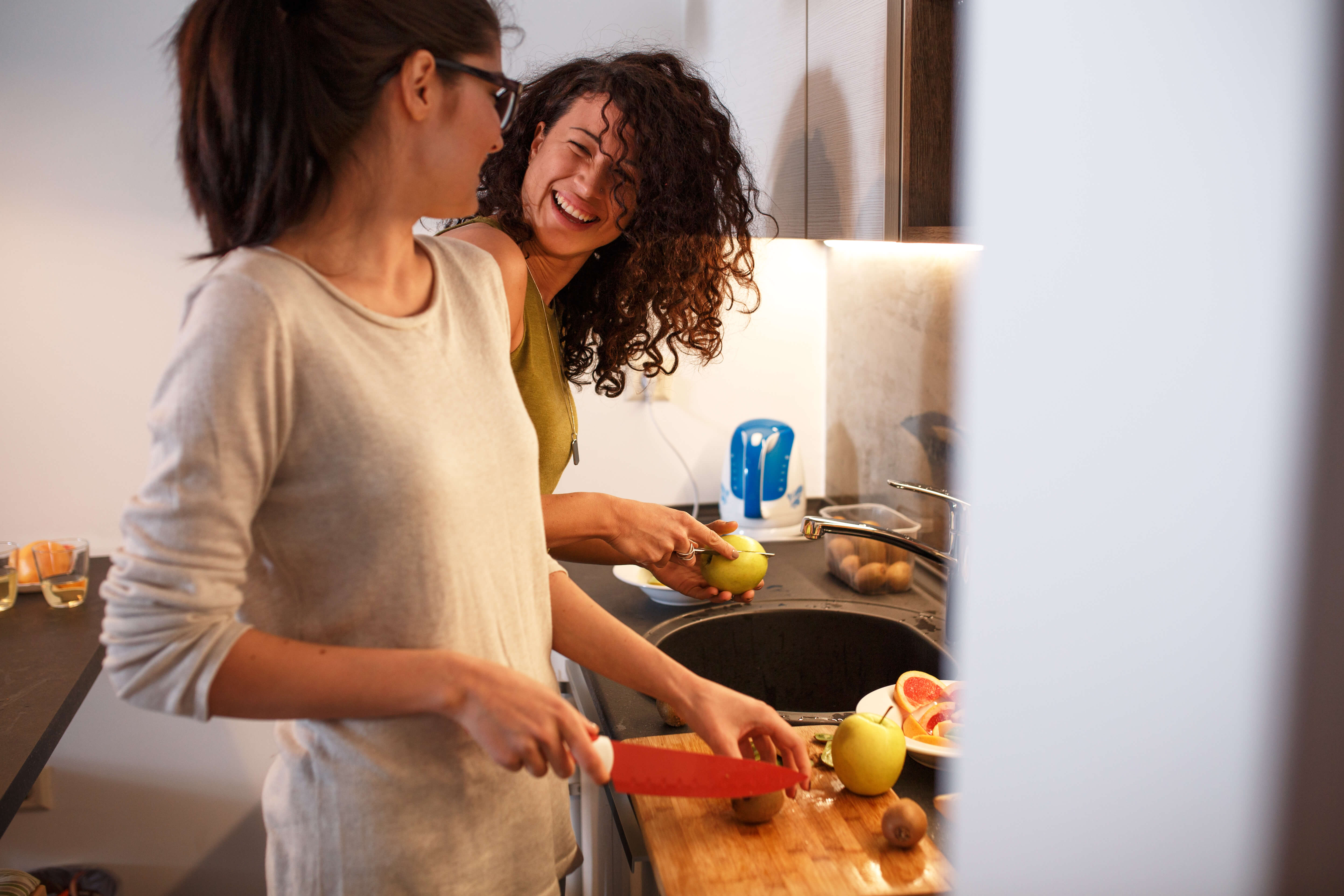 two women laughing and cutting fruit in a kitchen