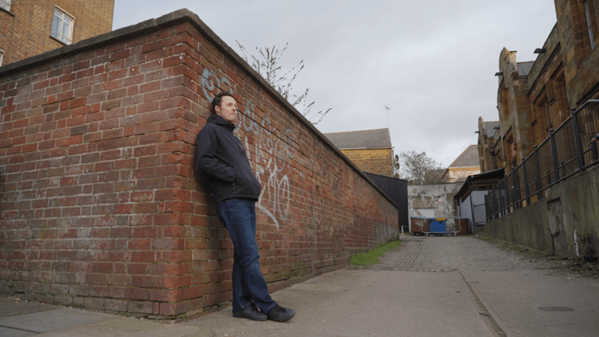 Full body view of a man named Kieran leaning against a wall with graffiti wearing a black jacket and blue jeans