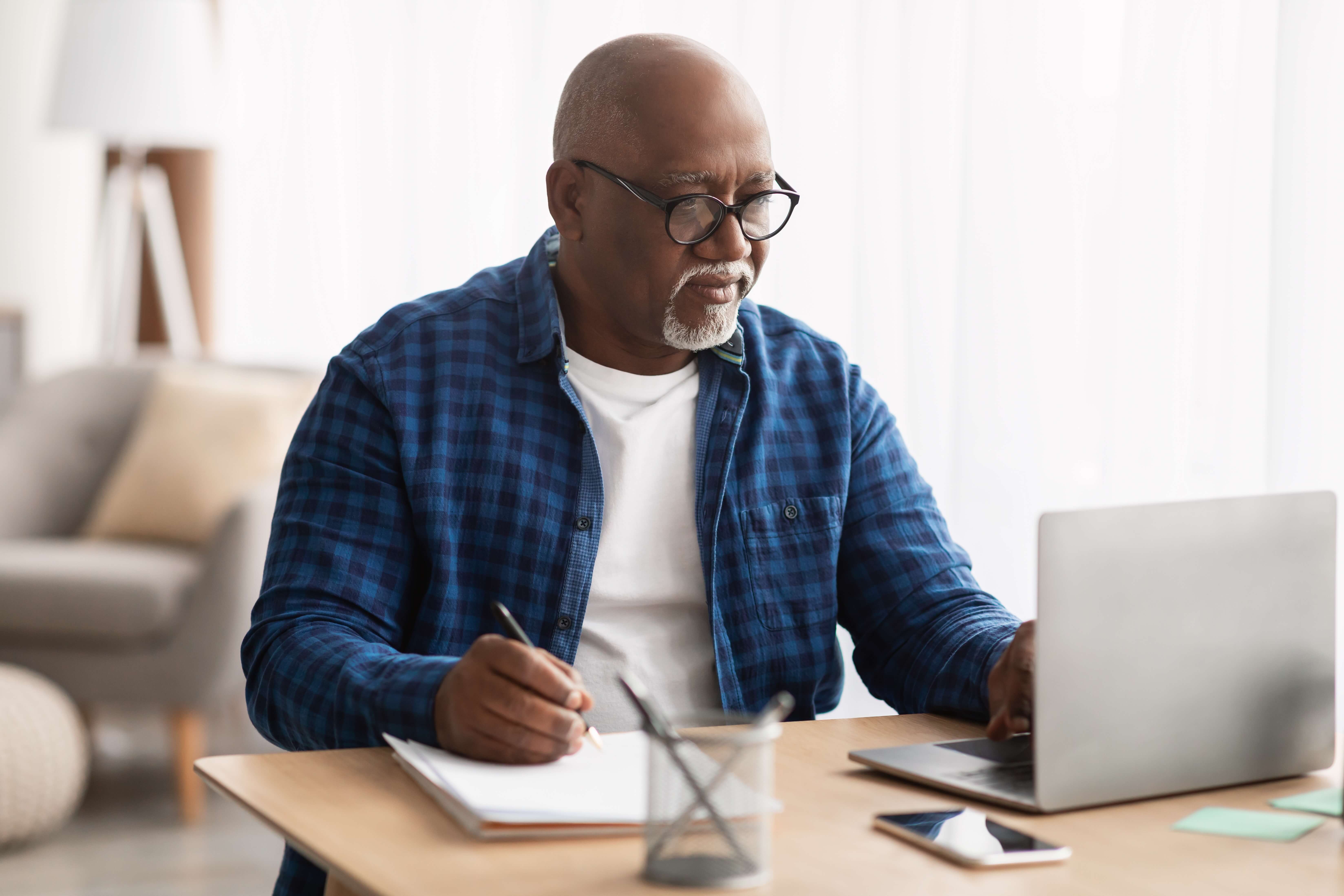 image of man in a blue shirt using a laptop and writing on a notepad
